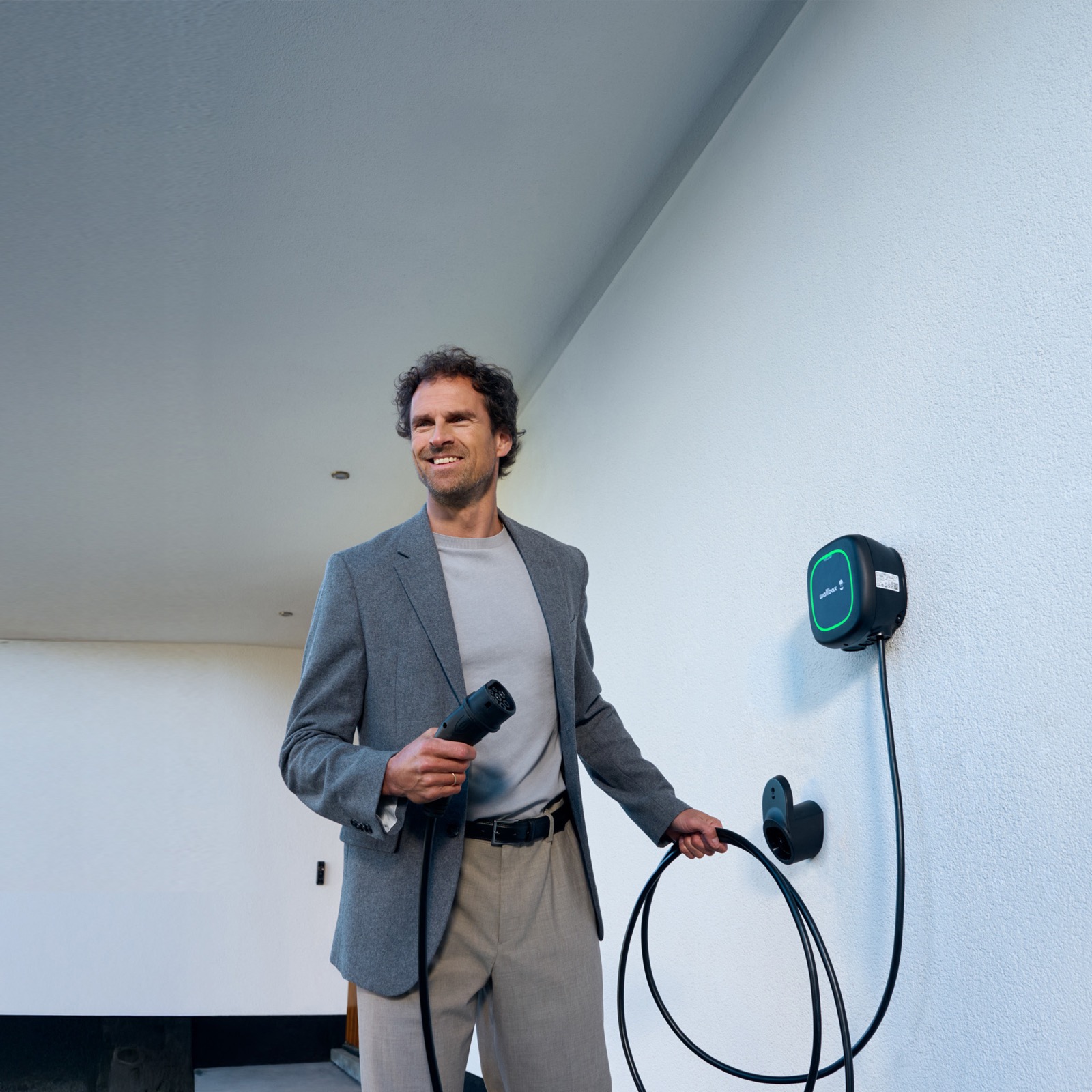Smilende mann med ladekabel ved en veggmontert Wallbox-lader hjemme